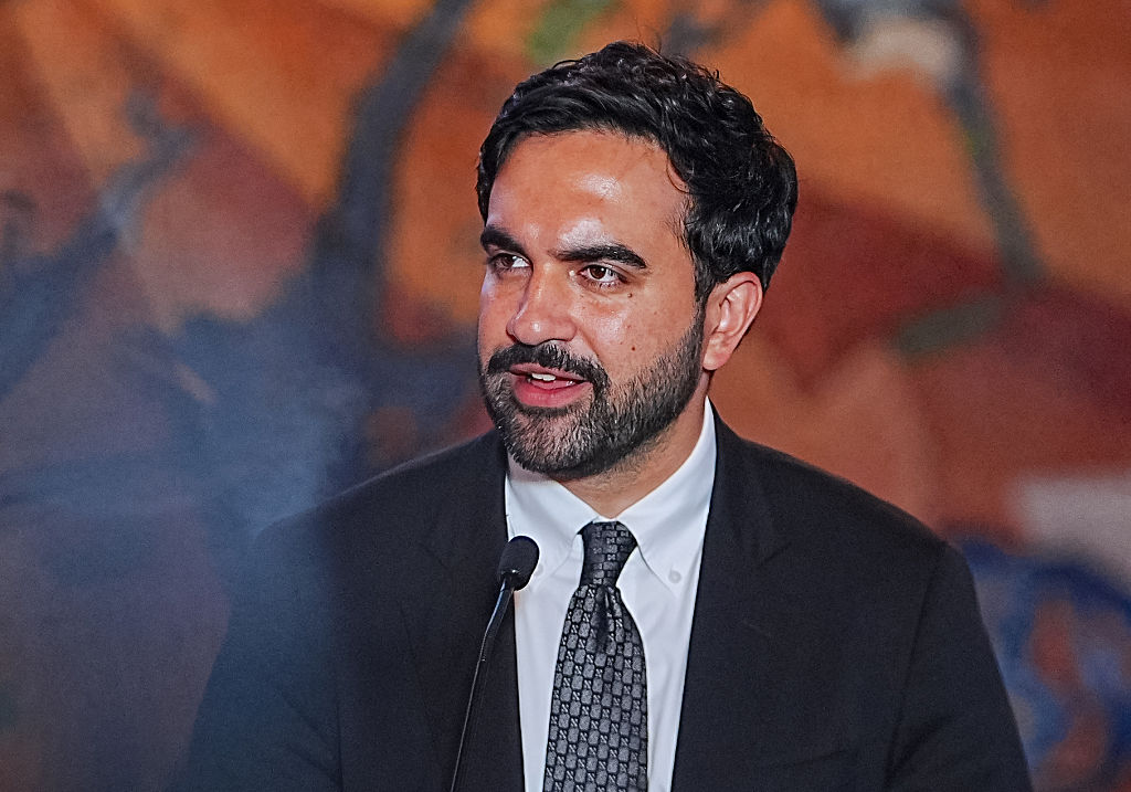 Mamdani, wearing a dark suit and patterned tie, speaks into a microphone against a blurred background.