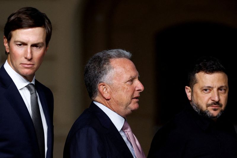 Three suited men stand together, including one wearing a black military-style shirt.
