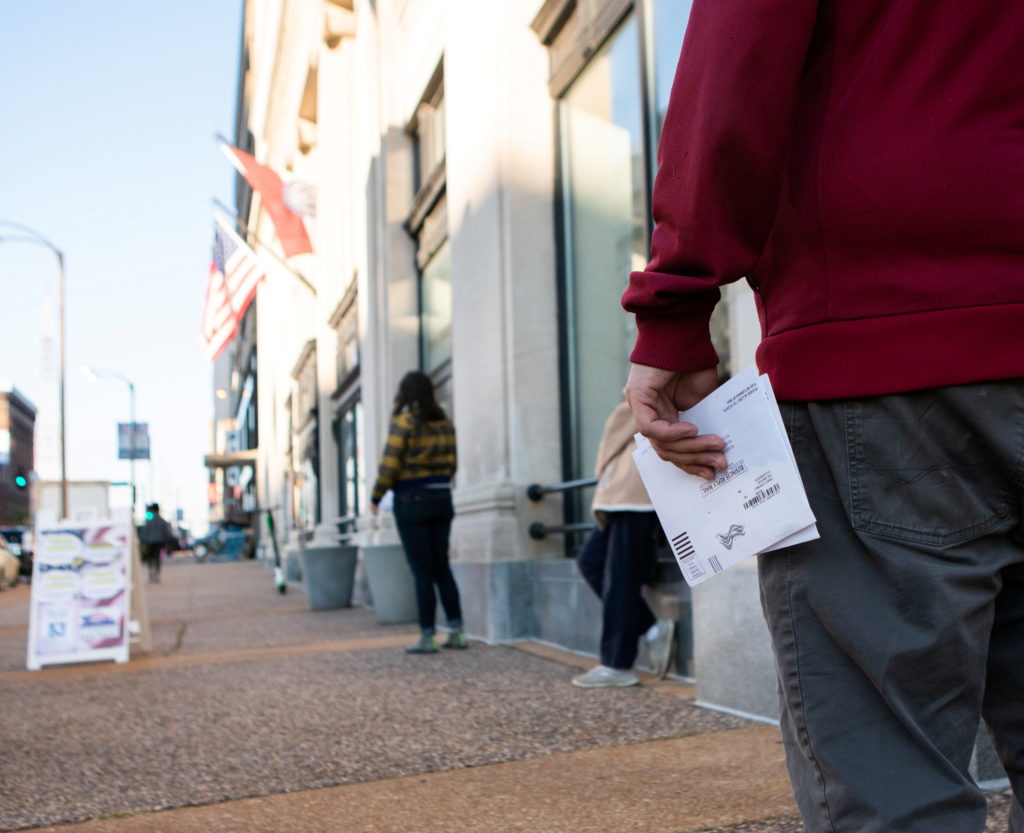 A voter holding an envelope waits in a line outside a building.