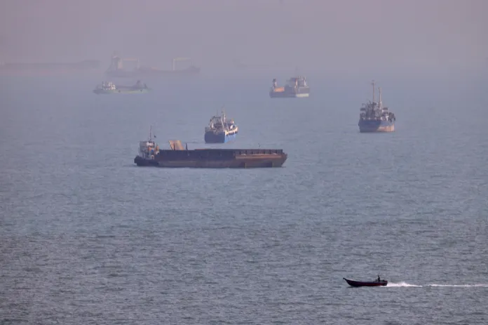 Several cargo vessels sit in hazy waters as a small motorboat crosses the foreground.