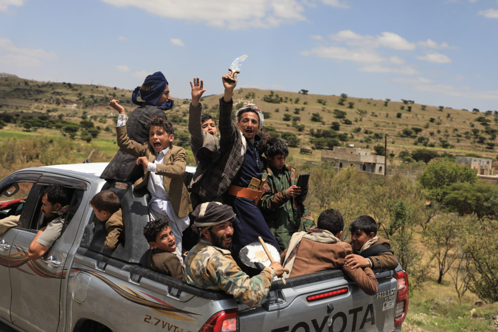 Men and boys ride in a pickup truck, with several raising their arms and one holding a curved blade.
