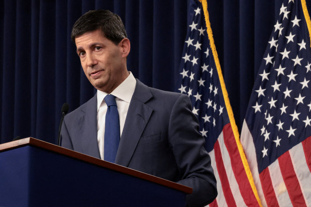Fed chair Kevin Warsh speaks from a lectern in front of dark curtains and two American flags.