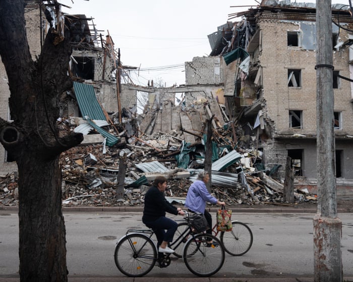 Two women ride bicycles past heavily damaged brick buildings and piles of rubble in Ukraine.
