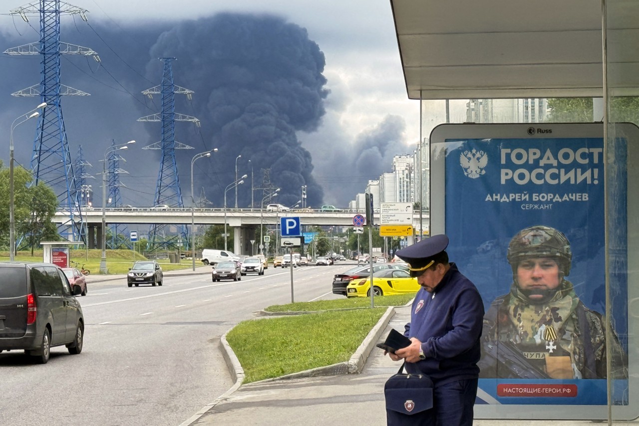 A vast dark smoke plume rises beyond power lines and city buildings near a bus shelter displaying a Russian military advertisement.