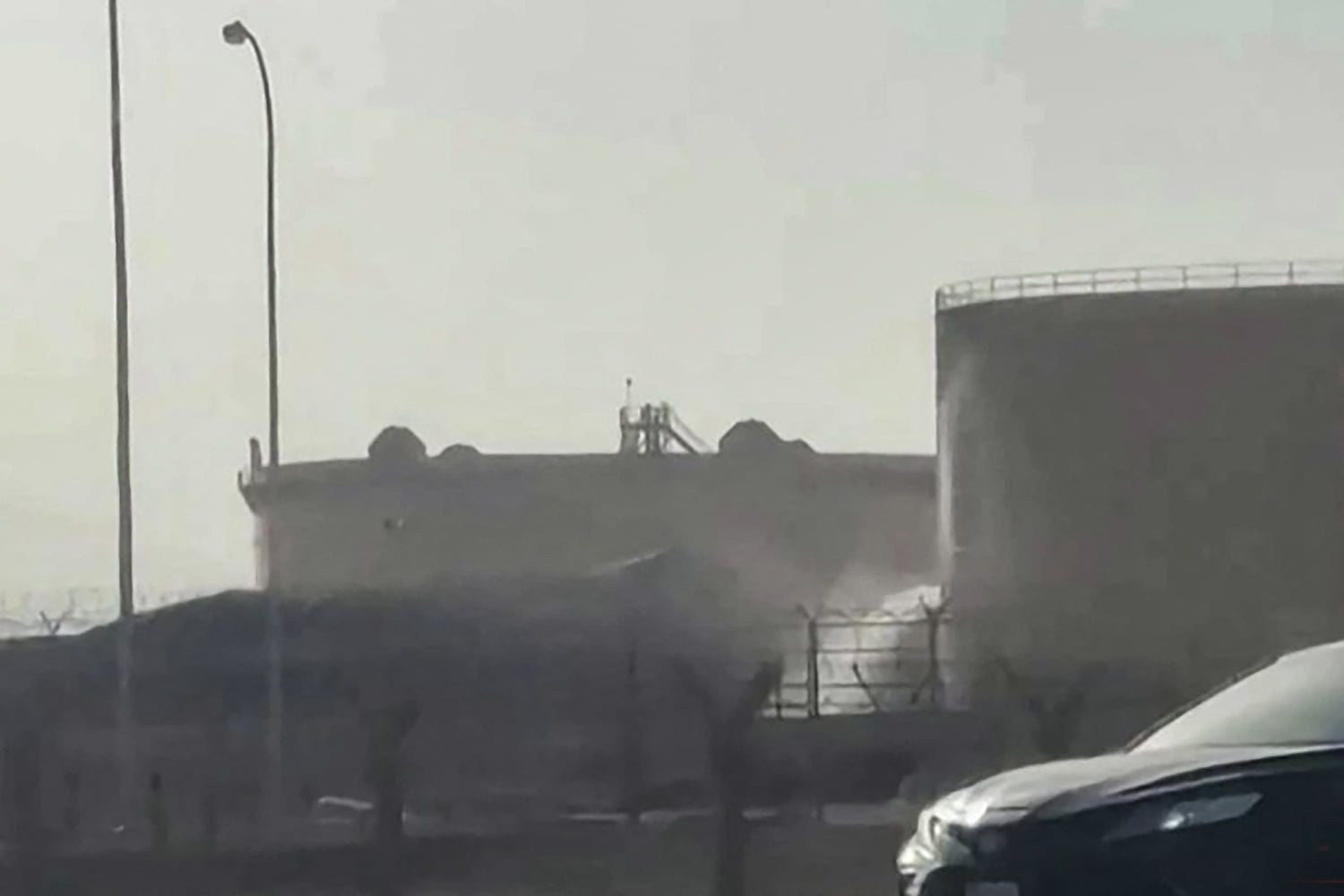 Smoke drifts over a fuel storage depot behind security fencing.