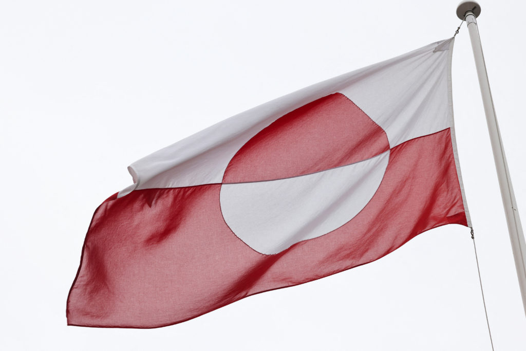 Greenland's red-and-white flag billows from a flagpole against a pale, overcast sky.