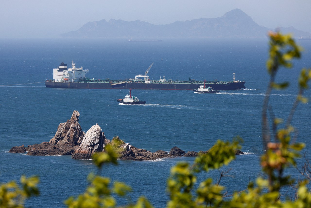A tanker under tow by tugboats passes rocky outcrops along a coast, with mountains in the haze.