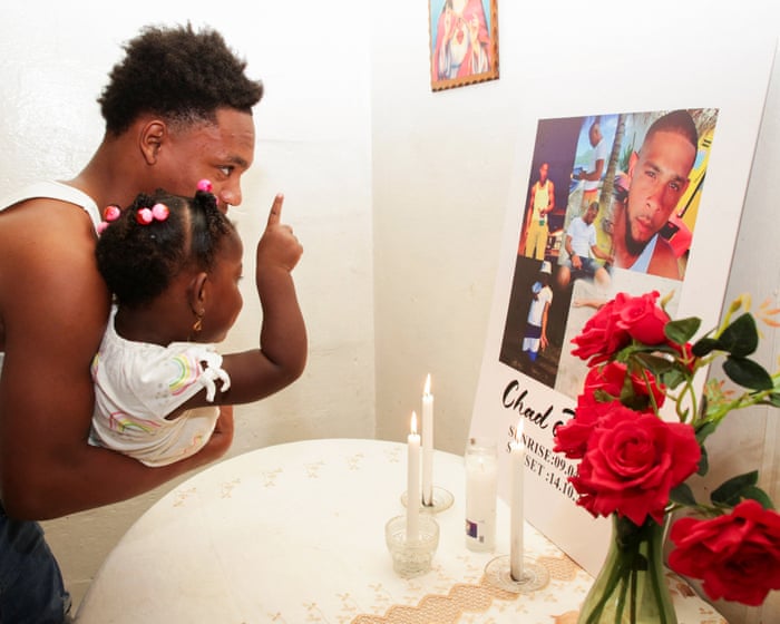 A man holding a young child gestures toward a candlelit memorial with red roses and a portrait board.