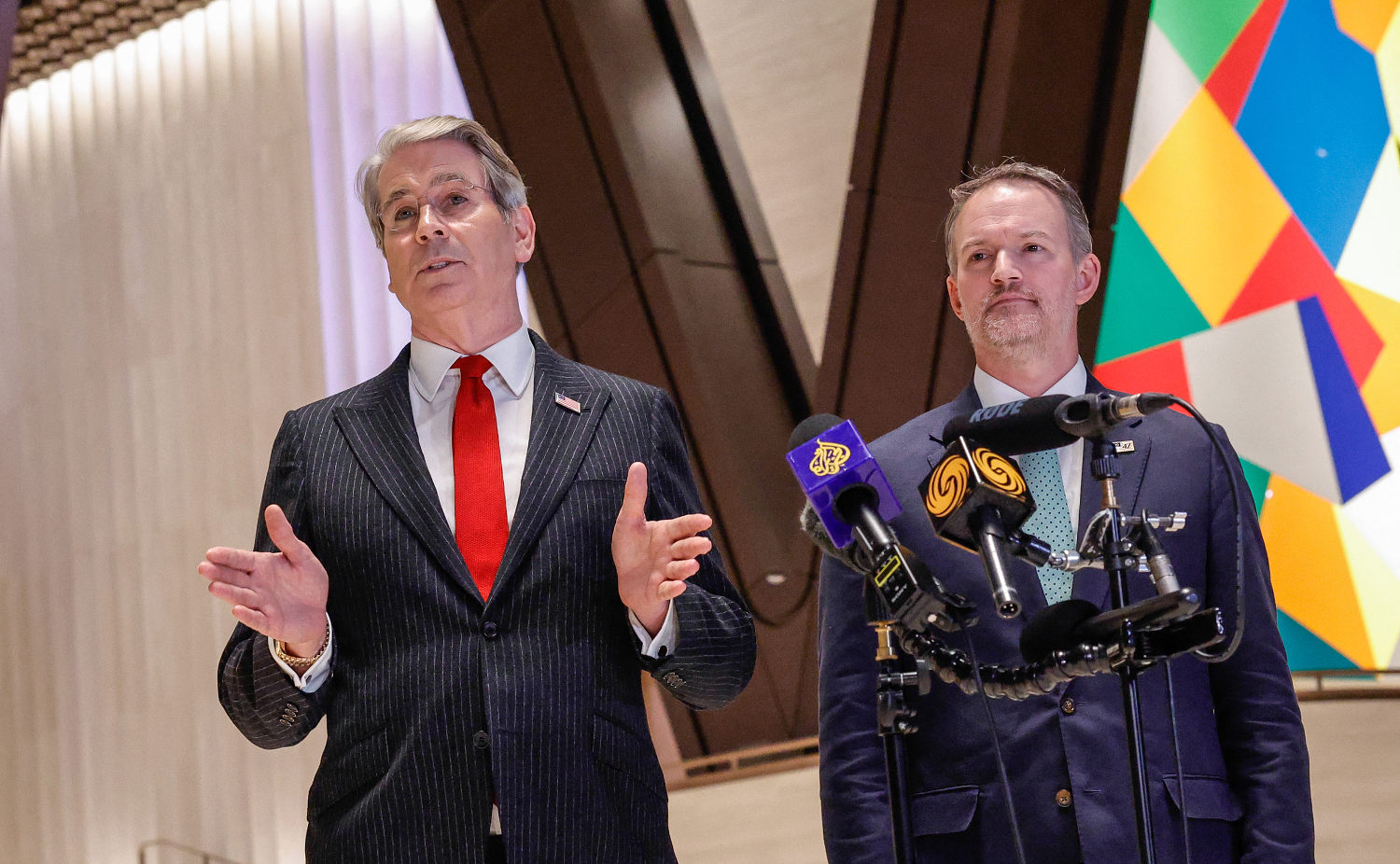 Two suited officials stand behind clustered microphones as one gestures during remarks.