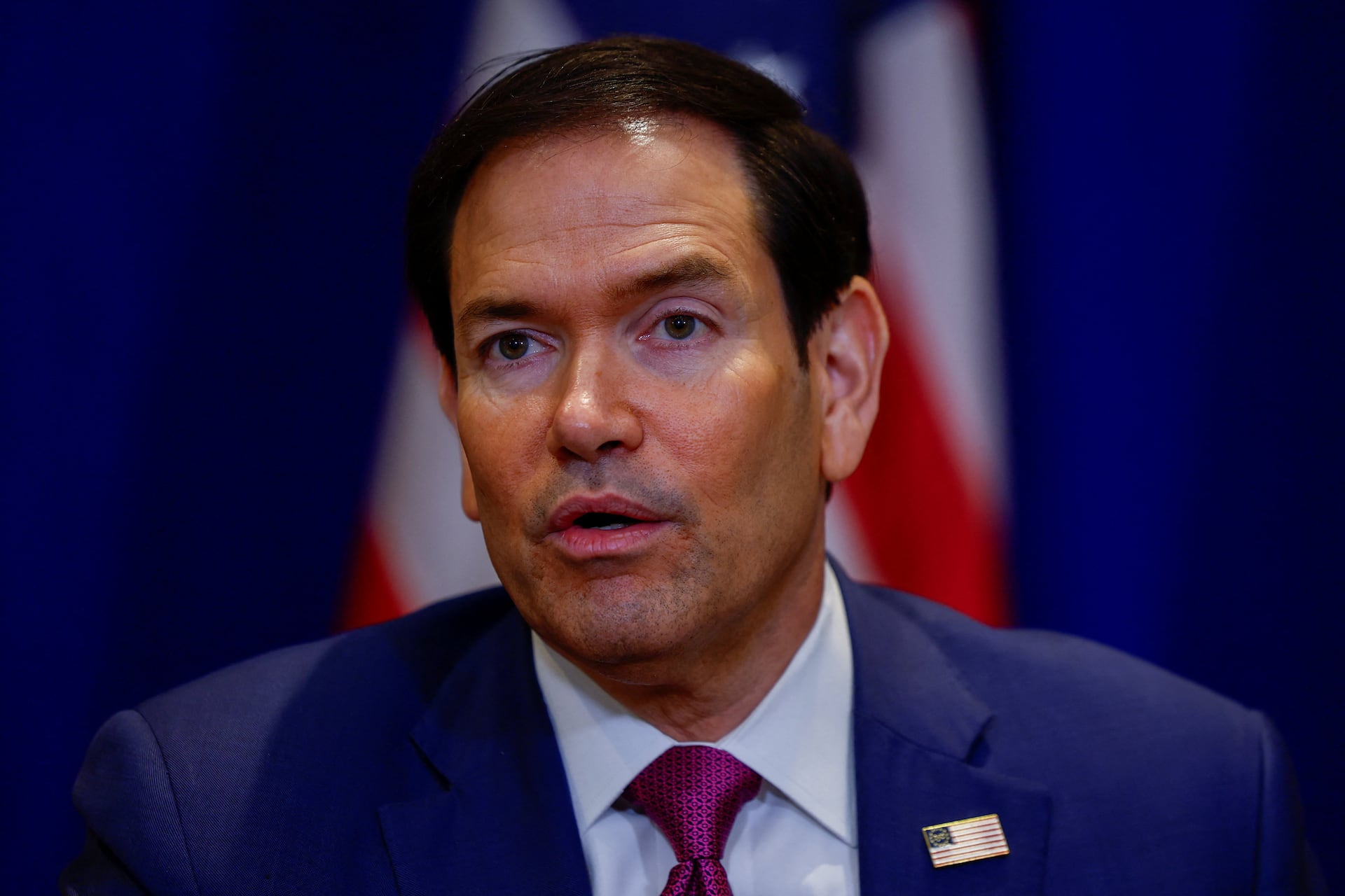 Rubio appears in close-up wearing a dark suit, patterned red tie, and American flag lapel pin.