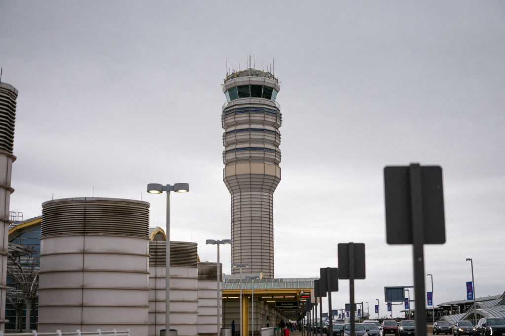 An airport control tower rises above terminal structures, roadway signs, streetlights, and vehicles beneath an overcast sky.