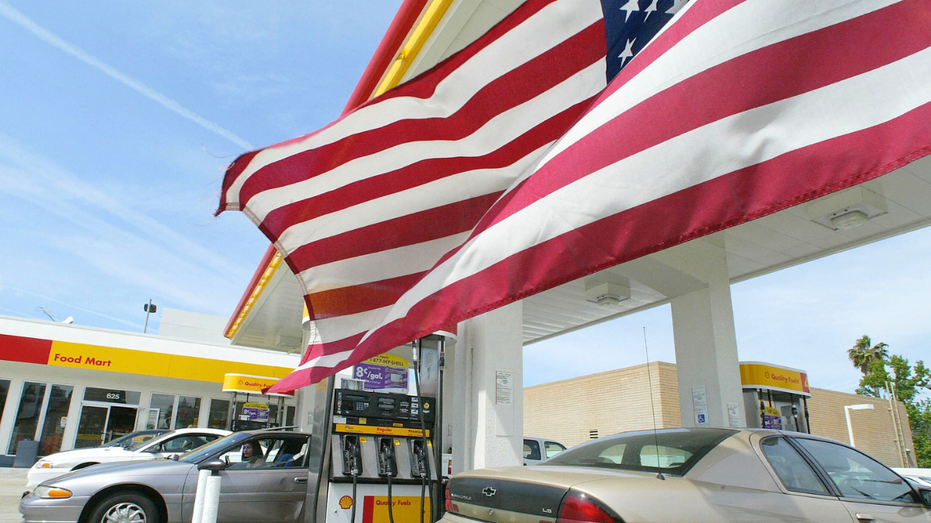 Two large American flags hang above fuel pumps and parked cars at a gas station.