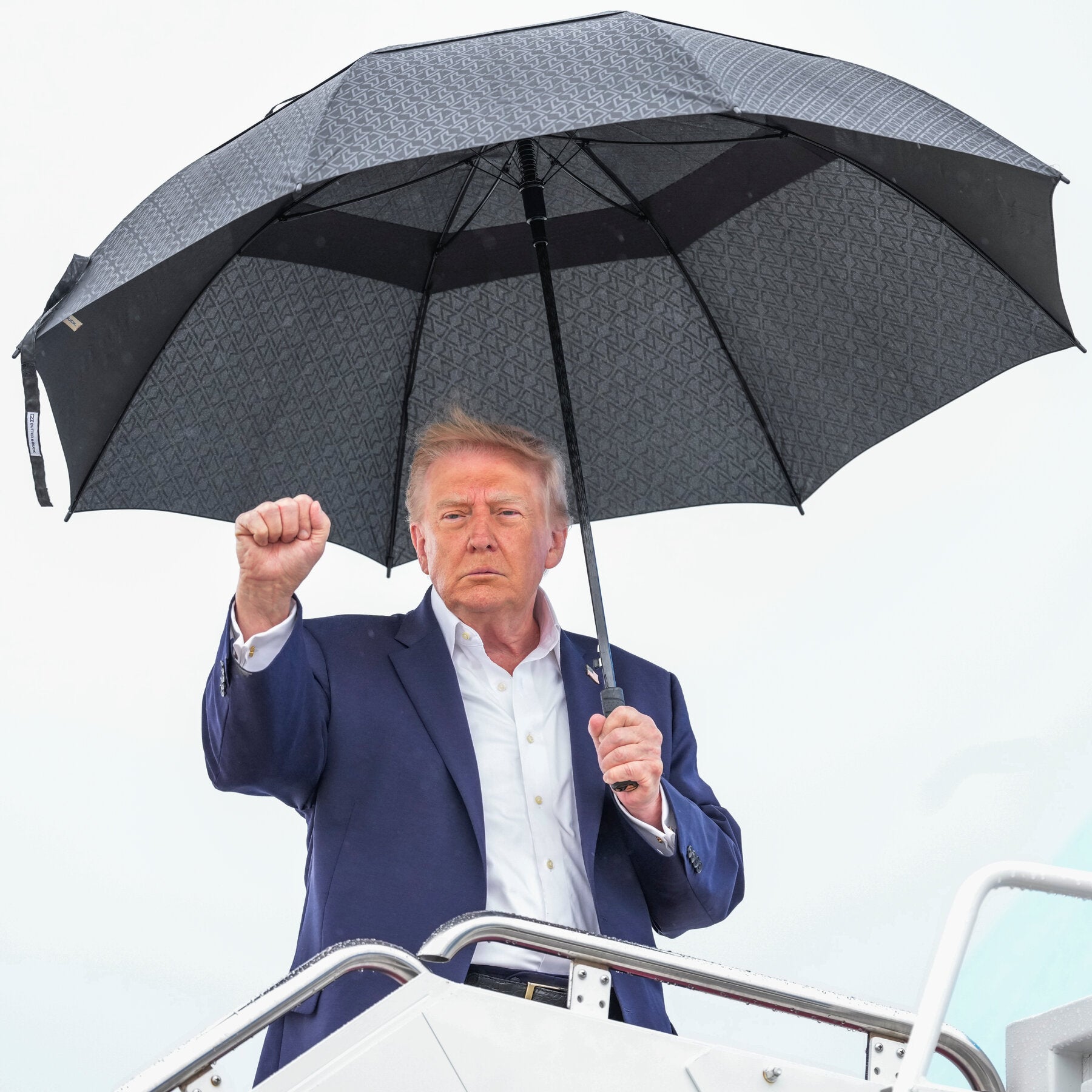 Trump raises a fist while holding a large umbrella on airplane stairs.