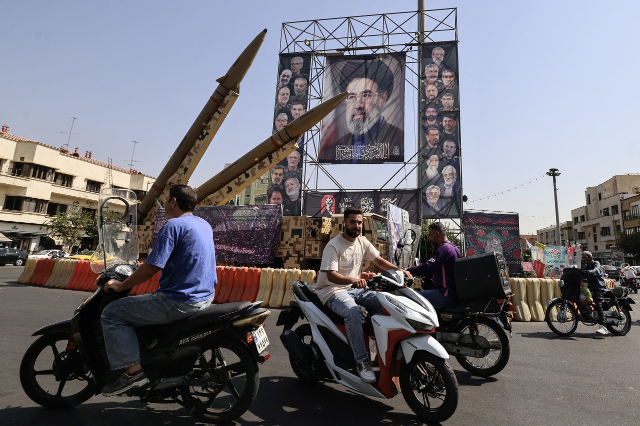 Motorcyclists ride past a street display of two missiles beneath a large portrait of a cleric on a city square.
