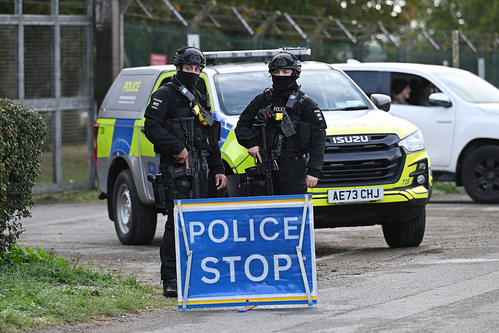 Two armed police officers in balaclavas stand behind a Police Stop sign, a marked police vehicle and security fencing behind them.