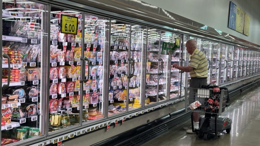An older shopper reaches into a refrigerated case in a long grocery aisle, a motorized cart beside him.