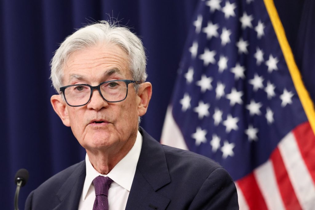 Powell, in glasses and a dark suit, speaks at a microphone in front of an American flag.