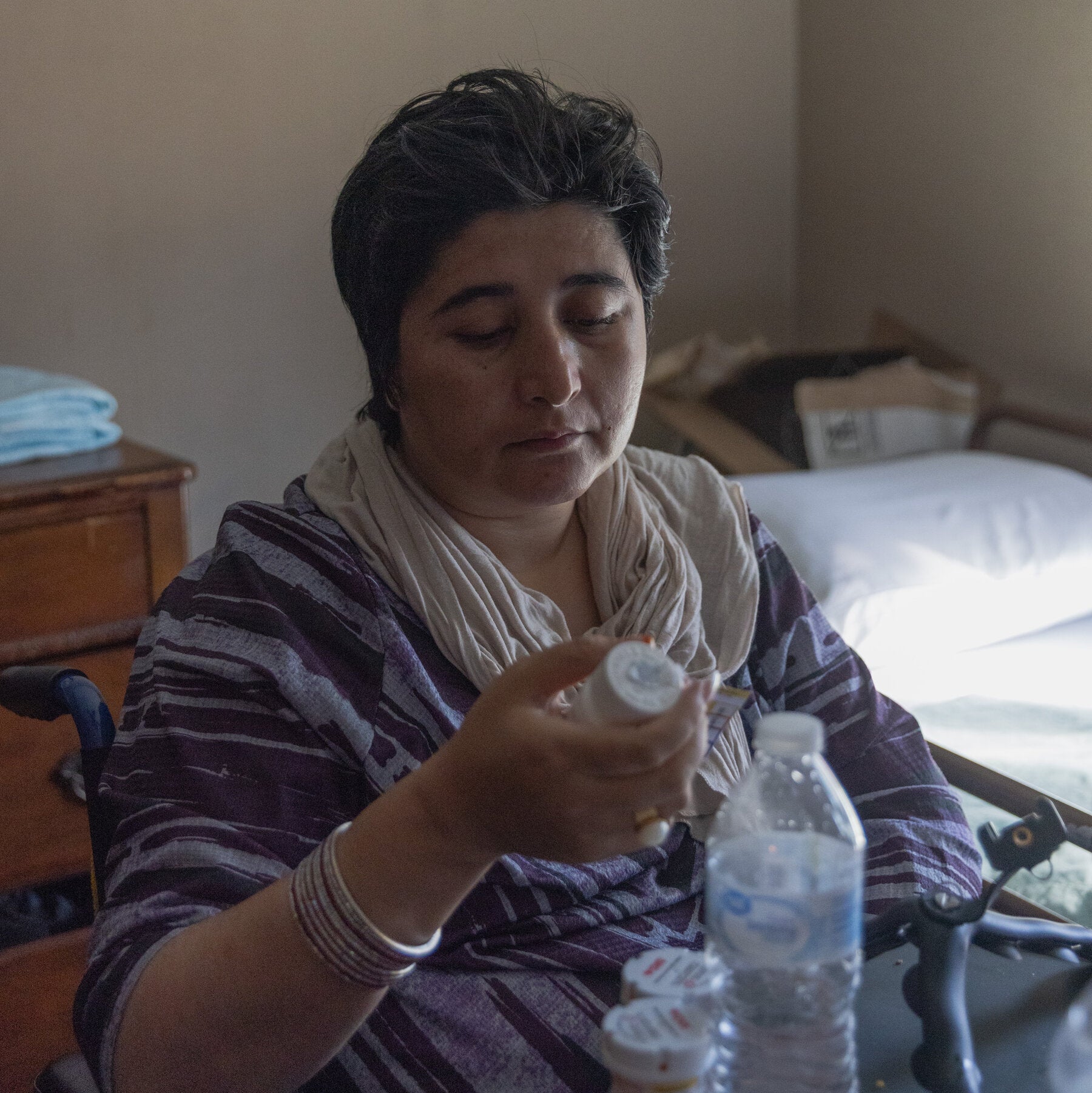 A seated woman examines a prescription bottle beside several medicine containers and a water bottle in a bedroom.