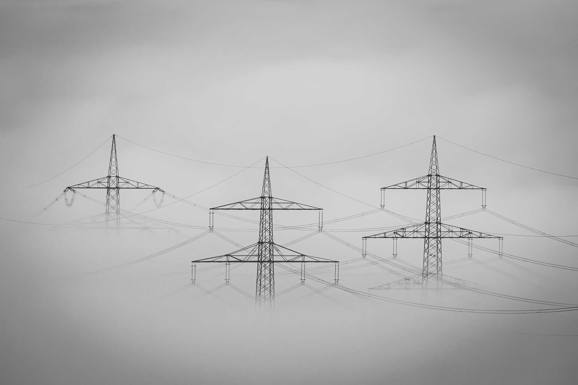 Three high-voltage transmission towers rise out of thick fog, their lines fading into gray, in a black-and-white photograph.