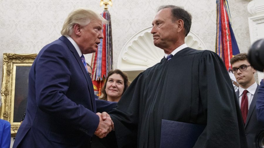 Trump shakes hands with Alito, who wears judicial robes, in a formal room with flags behind them.