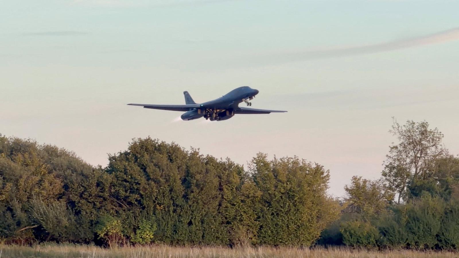 A B-1 bomber flies above a tree line with its landing gear lowered.
