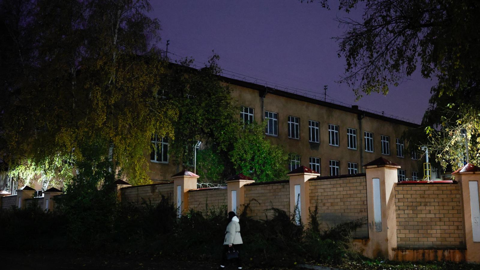 At dusk, a person in a light coat walks past a brick perimeter wall, with trees and a three-story building behind it.