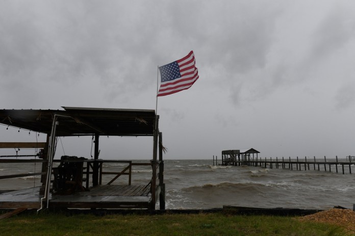 An American flag snaps in the wind above a wooden waterfront shelter as choppy gray waves roll past a long pier under a low, stormy sky.