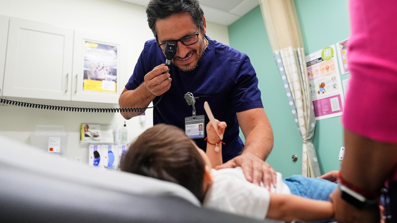 A clinician in navy scrubs smiles as he examines a young child lying on an exam table, holding an instrument to his eye.