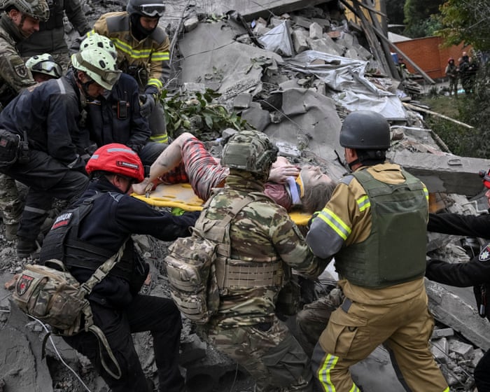 Rescuers in helmets and soldiers in camouflage carry an injured woman on a yellow stretcher across a pile of broken concrete.
