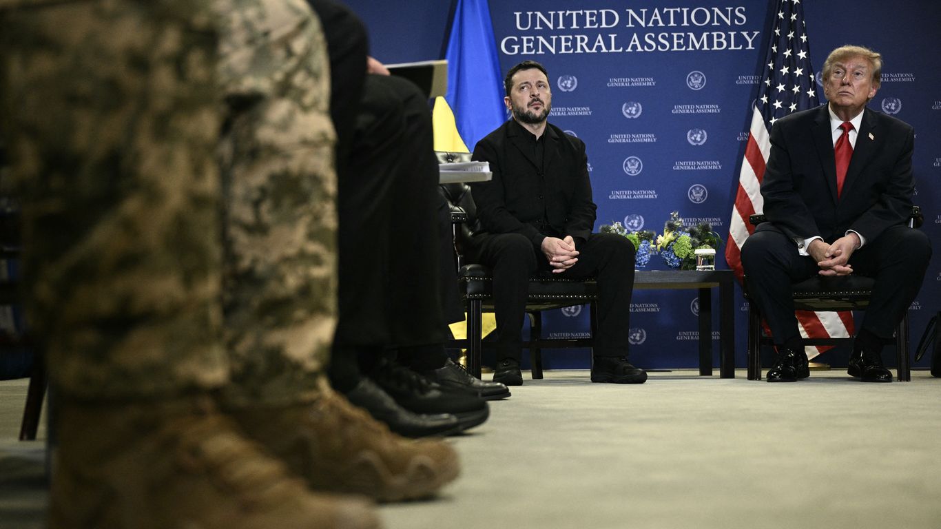 Volodymyr Zelensky and Donald Trump sit in chairs before a United Nations General Assembly backdrop, flags behind them, while soldiers’ legs and boots blur in the foreground.