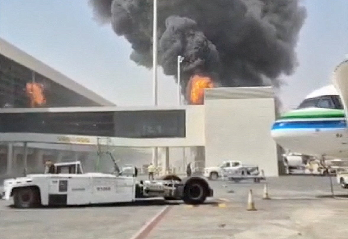 Black smoke and orange flames rise behind an airport terminal; a white aircraft tug sits on the apron, and the nose of a green-and-white airliner enters at the right edge.
