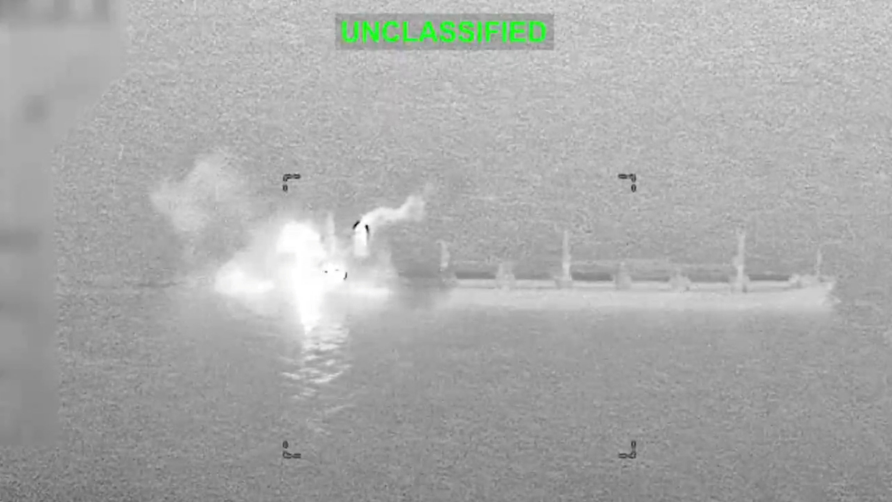 A grainy black-and-white infrared video frame marked “Unclassified” shows a bright burst and smoke at one end of a long cargo ship at sea.