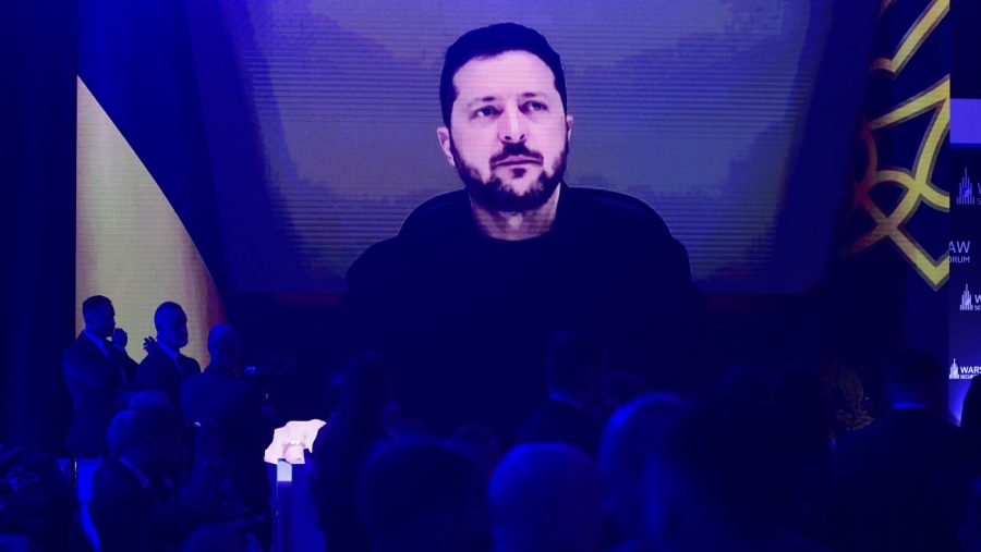 Volodymyr Zelensky appears on a giant screen above a blue-lit conference hall, the audience below in silhouette.