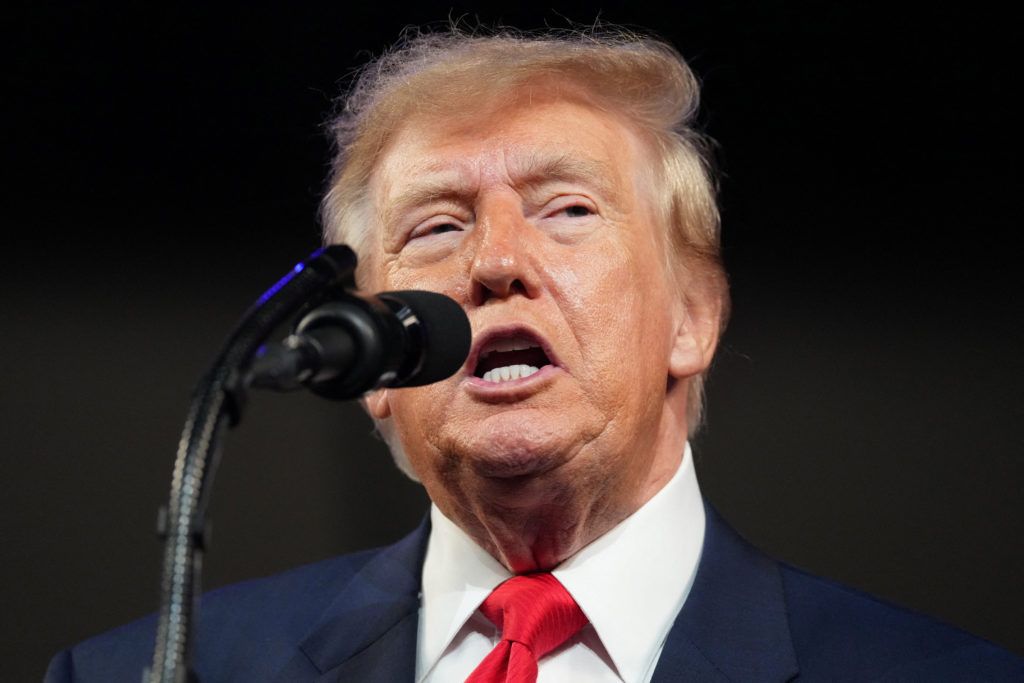 Trump, in a dark suit and red tie, speaks into a microphone against a black background.