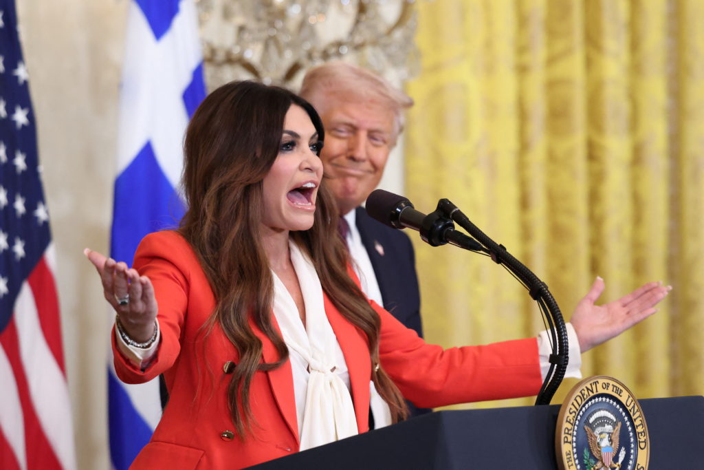 Kimberly Guilfoyle, in a red jacket, speaks with her arms spread at a lectern bearing the presidential seal as Donald Trump stands behind her, with U.S. and Greek flags at left.