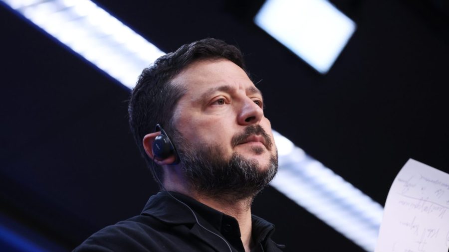 Volodymyr Zelensky, wearing an earpiece and a dark collared shirt, looks up past the camera, a handwritten sheet at the right edge.